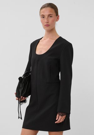 Femme portant une robe noire à manches longues avec des poignets en dentelle, tenant un sac noir à cordon sur un fond clair uni.