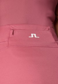 Růžový sportovní top s hladkým povrchem a přiléhavým střihem, se malou kapsou na zip a bílým logem vpředu.