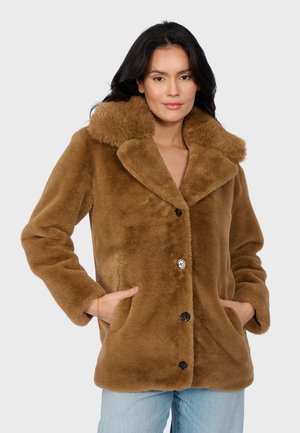 Donna che indossa un cappotto in pelliccia sintetica marrone con ampio colletto e bottoni neri, con le mani nelle tasche, abbinato a jeans azzurri chiari, su uno sfondo neutro.