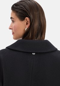 Zwarte wollen jas met een brede kraag, zichtbare naaddetails en een decoratieve metalen knoop aan de achterkant. Gladde textuur en getailleerd ontwerp.