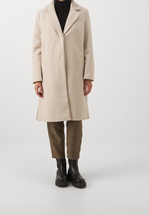 Personne portant un manteau beige mi-long sur un pantalon marron à carreaux et des bottines noires épaisses, debout devant un fond clair uni.