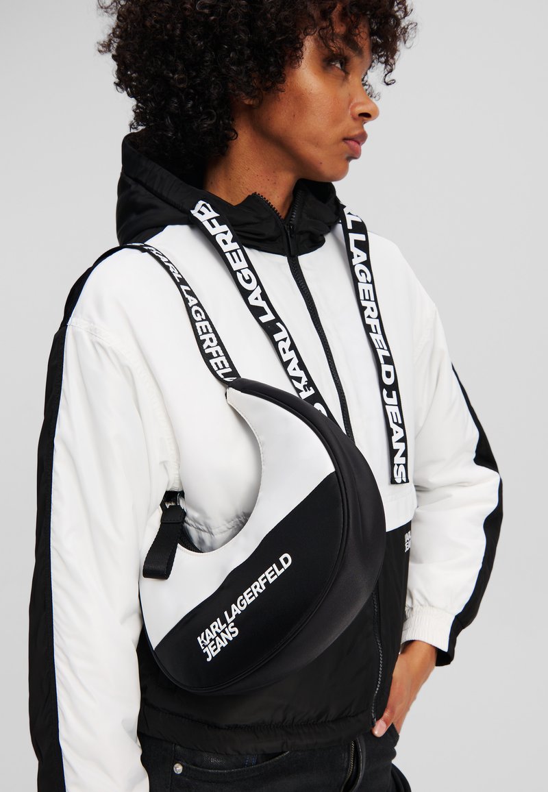 Juoda ir balta petneša formos kuprinė, pasižyminti lygiu audiniu, logotipo detalėmis ir reguliuojama dirželiu.