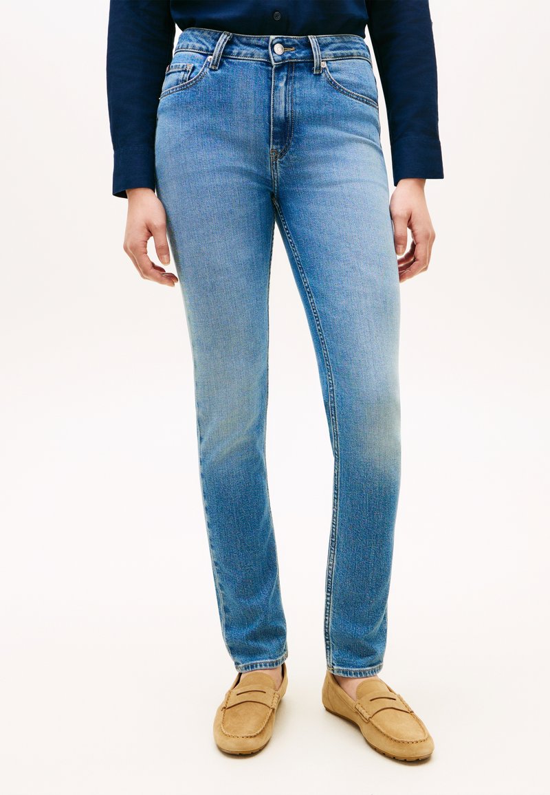 Άτομο που φοράει στενά τζιν ανοιχτού μπλε χρώματος, ταμπά μοκα σίνια και σκούρο μπλε πουκάμισο με μακριά μανίκια, στέκεται μπροστά σε απλό φόντο.