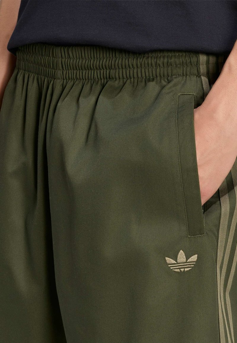 Primer plano de una persona que lleva pantalones deportivos Adidas verde oliva con cintura elástica, mano en el bolsillo y visible el logo de trébol de Adidas.