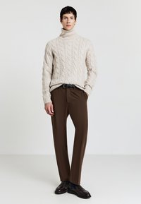 Beige Kabelstrick-Turtleneck-Pullover, braune maßgeschneiderte Hose, schwarzer Gürtel und dunkle Schuhe. Glatte Textur und harmonisches Farbschema.