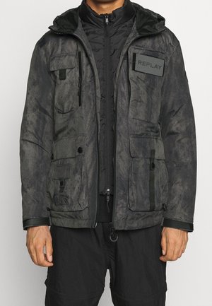 Mężczyzna w ciemnoszarej kurtce z kapturem, z wieloma kieszeniami z przodu i naszywką "REPLAY" na czarnej, rozpinanej wewnętrznej warstwie.