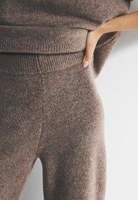 Zbliżenie osoby noszącej dopasowany brązowy sweter i spodnie z dzianiny, z jedną ręką opartą na talii na jednolitym białym tle.