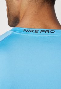 Nike Pro Sporthemd in Hellblau mit glatter Textur und schwarzem Logo am Ausschnitt. Nähte sind entlang der Ränder sichtbar.