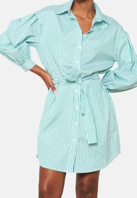 Koszula sukienkowa w jasnym odcieniu teal z białymi paskami, z długimi bufiastymi rękawami oraz przewiązywanym paskiem w talii dla podkreślenia kształtu.