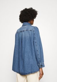 Camisa de denim de corte relajado, de color azul oscuro, con cuello, puños abotonados y un diseño con pliegues en la parte posterior, mostrada desde la parte trasera.