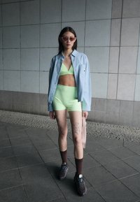 Ensemble de maillot de bain à deux pièces vert néon complété par une chemise boutonnée bleu clair, des chaussettes brunes courtes et des baskets sombres ; tatouages visibles sur les jambes.
