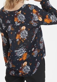 Top à manches longues en tissu foncé avec des motifs floraux et de papillons en orange, gris et blanc. Il présente une coupe décontractée et un col rond.