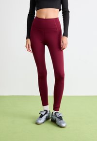 Mörkröda högmidjade leggings av elastiskt tyg, med en slät yta och subtil sömdetalj, kombinerat med grå träningsskor.
