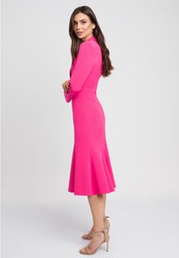 Dopasowana, jasnoróżowa sukienka z długimi rękawami i rozkloszowanym dołem, zestawiona z cielistymi sandałami na wysokim obcasie. Gładka, bezszwowa faktura materiału.