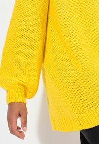 Maglione giallo a maglia con vestibilità ampia, polsi a coste e superficie testurizzata. Il tessuto appare morbido e voluminoso, offrendo calore.