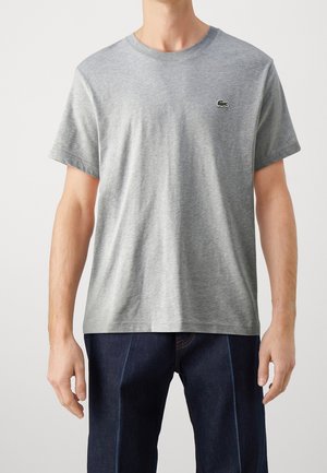 Mann trägt ein schlichtes graues T-Shirt mit kurzen Ärmeln und kleinem grünem Krokodil-Logo auf der Brust sowie dunkelblaue Jeans.