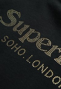 Tessuto nero con la scritta "Superdry" impreziosita da strass dorati, superficie testurizzata e finitura liscia.