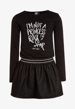 Czarna bluzka z długim rękawem z białym nadrukowanym tekstem; czarna spódnica z błyszczącego materiału z srebrnym akcentem w pasie. Oba elementy wykonane są z miękkiego materiału.