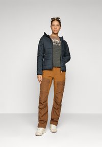 Doudoune bleu foncé, pull à motifs dans des tons terreux, et pantalon cargo marron avec poches, porté avec des baskets de couleur claire.