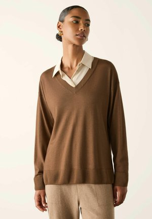 Kvinde iført en brun V-hals sweater over en cremefarvet skjorte med krave og beige bukser, poserer mod en ensartet lys baggrund.