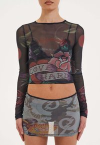 Durchsichtige schwarze Langarmbluse mit floralen Tattoos und dem Aufdruck "LOVE HARD"; kombiniert mit einem halbtransparenten Rock, der verschiedene Muster und Farben aufweist.
