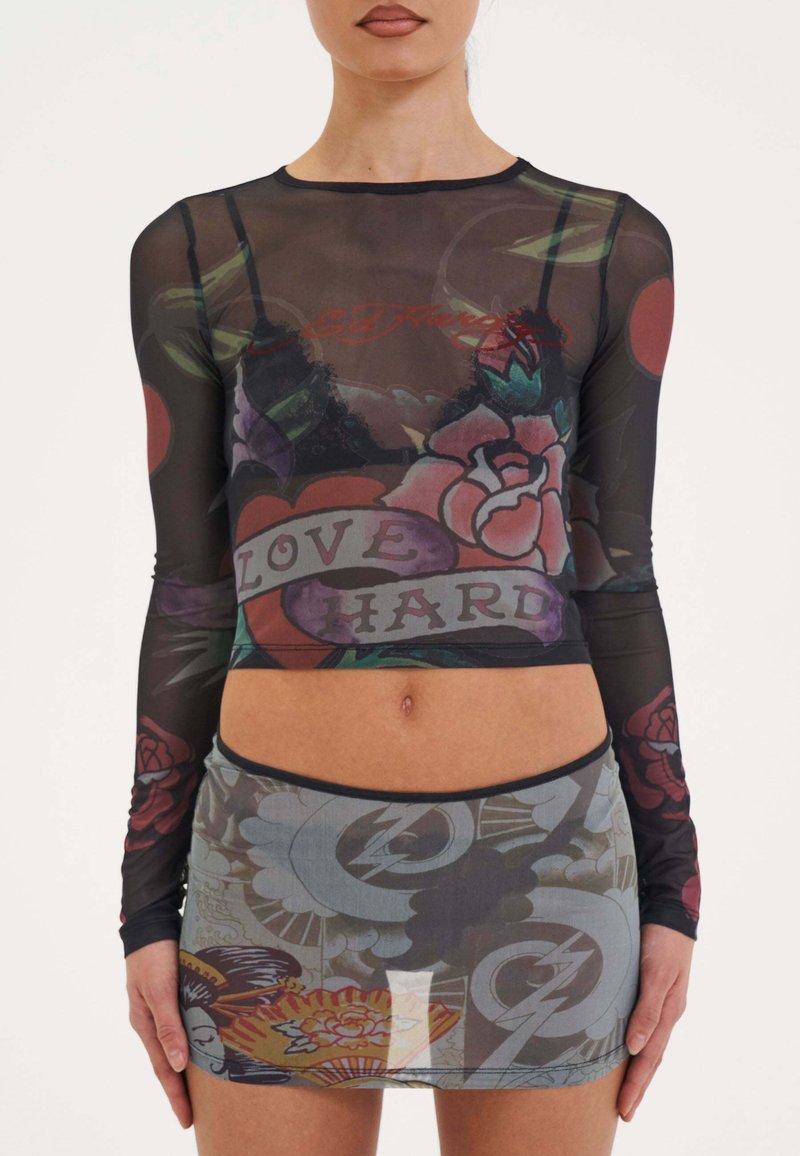 Durchsichtige schwarze Langarmbluse mit floralen Tattoos und dem Aufdruck "LOVE HARD"; kombiniert mit einem halbtransparenten Rock, der verschiedene Muster und Farben aufweist.