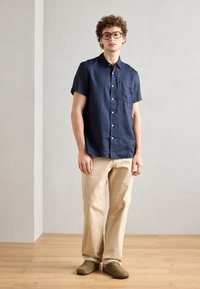 Marineblauw linnen overhemd met korte mouwen en een knoopsluiting, beige wijde broek, en bruine instap schoenen op een houten vloer.