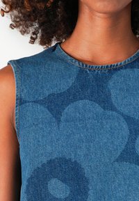 Denim-ermløs topp med blå teksturert stoff og et subtilt mønster av sirkulære design. Avsluttet med en enkel rund hals og sømmer.