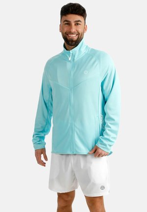 Mann lächelt, trägt eine hellblaue Jacke mit Reißverschluss und weiße Shorts, steht vor einem schlichten hellgrauen Hintergrund.