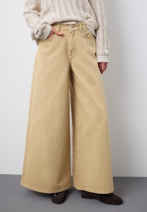 Person trägt beige Hose mit weitem Bein, cremefarbenen Strickpullover mit den Händen in den Taschen und dunkelbraune Mokassins und steht vor einem einfarbigen Hintergrund.