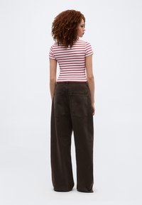 Γυναίκα με σγουρά μαλλιά στέκεται γυρισμένη πλάτη, φορώντας εφαρμοστή κοντομάνικη μπλούζα με λευκές και κόκκινες ρίγες και πλατιά σκούρα καφέ παντελόνια.