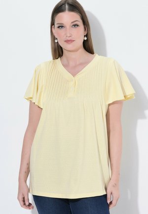 Kvinde iført en gul kortærmet bluse med plisseret V-halsudskæring og knapper, kombineret med mørkeblå jeans og perleøreringe.