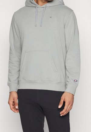 Man draagt lichtgrijze Champion-hoodie met voorzak en trekkoordjes, gecombineerd met zwarte broek, tegen een effen achtergrond.