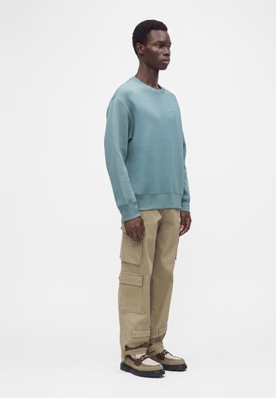 Homme debout portant un sweat-shirt bleu à manches longues, un pantalon cargo beige et des mocassins beige et marron devant un fond blanc uni.