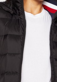 Doudoune noire avec capuche, dotée d'une fermeture éclair à l'avant et d'un design matelassé. En dessous, un t-shirt blanc uni.