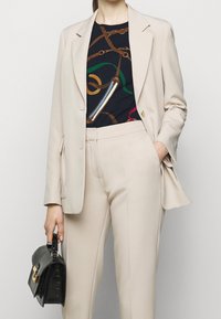 Beige skräddarsydd kavaj med skarpa slag, matchad med åtsittande byxor. Under kavajen en mörk mönstrad topp med guldfärgade och färgglada accenter. Svart strukturerad handväska.