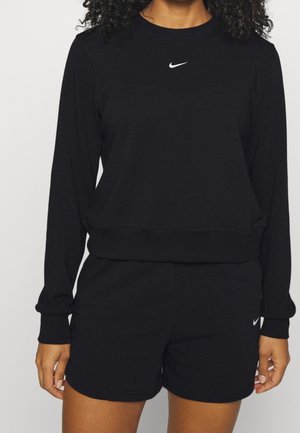 Personne portant un sweat-shirt noir à manches longues et un short avec des logos Nike blancs sur la poitrine et la cuisse gauche, debout devant un fond uni.