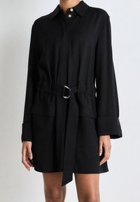 Abito camicia nero a maniche lunghe con cintura, dotato di due tasche frontali e polsini arrotolati, indossato da una persona in piedi davanti a uno sfondo neutro.