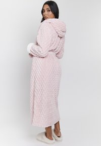 Accappatoio rosa, con texture, dotato di cappuccio e vita con cintura. Presenta polsini bianchi e un design lungo e fluente. Tessuto morbido con un aspetto accogliente.