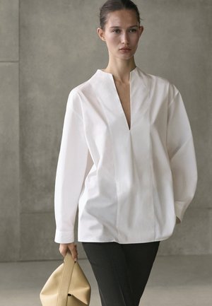 Kobieta idąca, ubrana w luźną białą bluzkę i czarne spodnie, trzymająca beżową, miękką torebkę o strukturze against na jednolitym szarym tle.