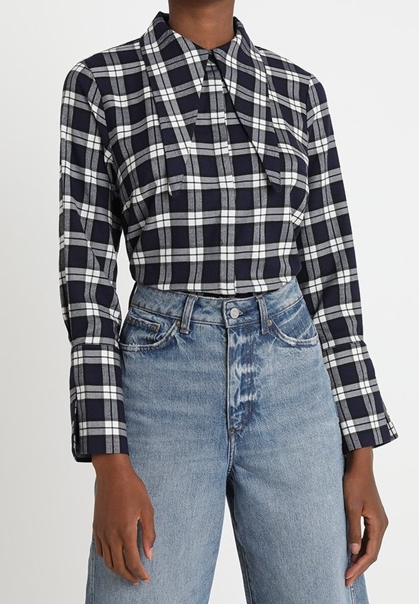Femme portant une chemise à carreaux noir et blanc à manches longues rentrée dans un jean en denim bleu clair taille haute.