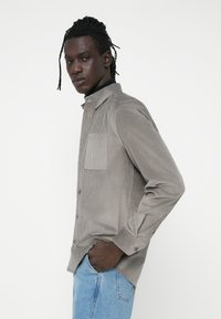 Camisa de pana gris con botones, con un bolsillo en el pecho, mangas largas y un cuello clásico. Llevada sobre un suéter de cuello alto azul marino y jeans azules.