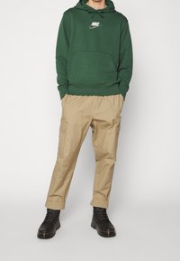 Grön hoodie med känguruficka och vit Nike-logga, kombinerad med beige cargobyxor och svarta snörskor. Enkel design; bomullsmaterial.