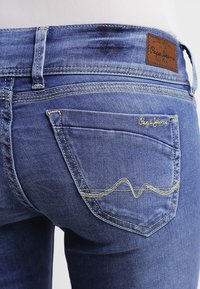 Blå denimjeans med en distinkt bakfickdesign, guldstickning och en läderlogotyp på midjan. Texturerad, åtsittande design.