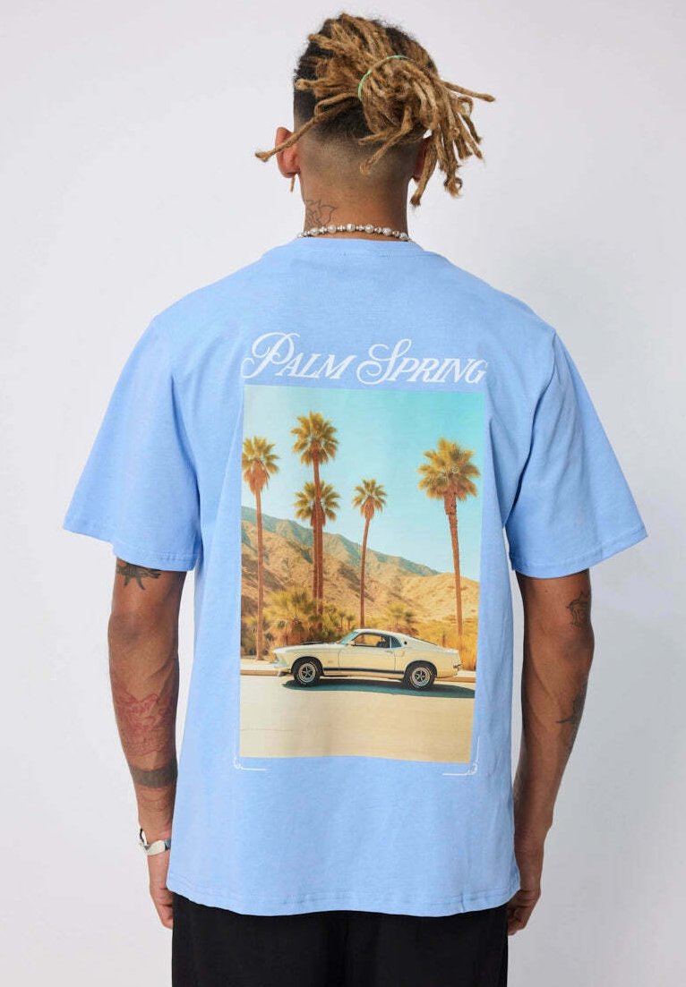 Personne avec des dreadlocks porte un T-shirt bleu clair affichant une voiture vintage et des palmiers sous le texte "Palm Spring" au dos.