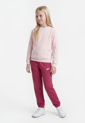 Ung pige stående iført en lyserød sweatshirt, mørkerøde Nike træningsbukser og hvide sneakers mod en hvid baggrund.