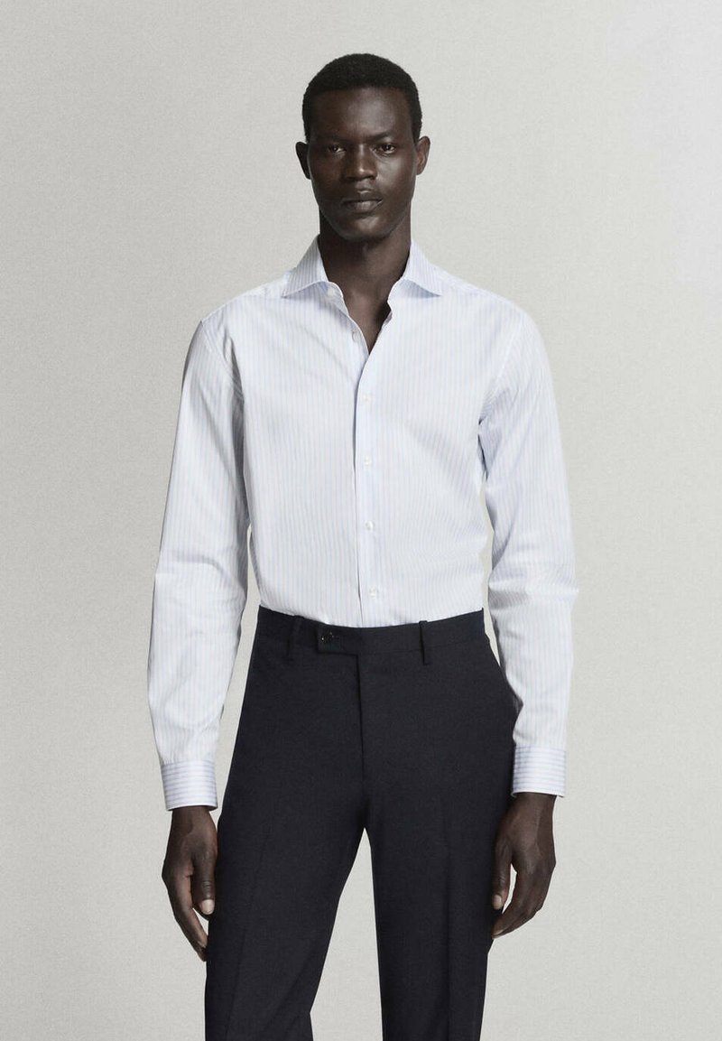 Homme portant une chemise à rayures bleu clair rentrée dans un pantalon noir sur mesure, sur un fond clair uni.