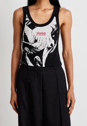 Kobieta ubrana w czarny top bez rękawów z białym motywem łabędzia i czerwonym napisem "HUGO LES BENJAMINS", w połączeniu z czarnymi spodniami z wysokim stanem.