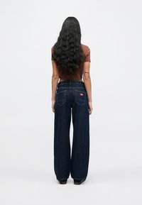 Γυναίκα με μακριά κυματιστά μαλλιά, ντυμένη με καφέ κοντομάνικο crop top, φαρδιά σκουρομπλε τζιν και μαύρα παπούτσια, στέκεται με την πλάτη στραμμένη.