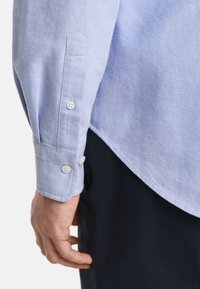 Hellblaues Hemd mit langen Ärmeln, ausgestattet mit weißen Knöpfen und gesticktem GANT-Logo am Manschette. Weicher Stoff, taillierte Passform.